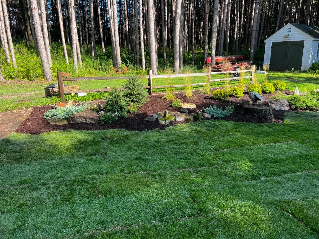 Lush garden landscape with stone features, plants, and wooden fence in a forest setting.