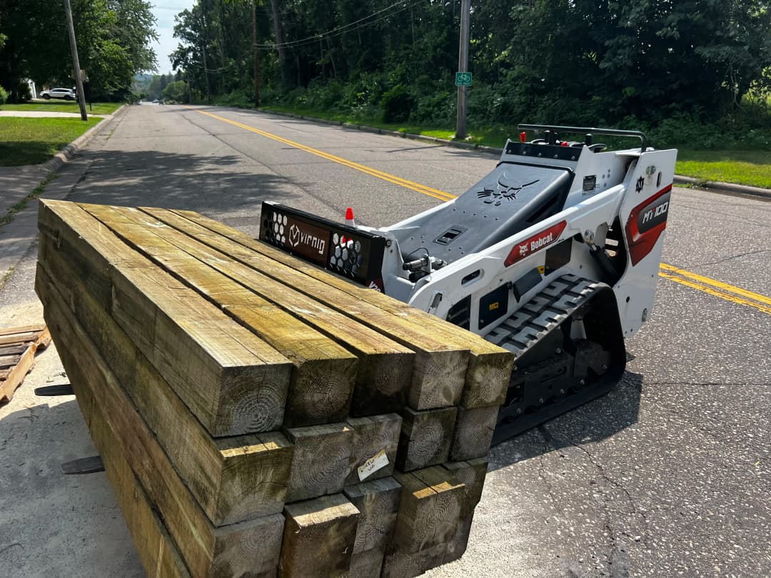 Skid steer loader beside stacked wooden beams on a suburban road.
