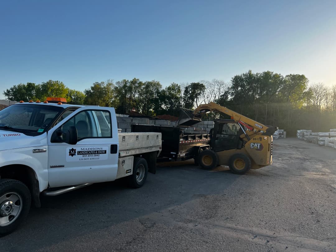 Pickup truck and skid loader at a construction site, loading materials with a clear sky background.