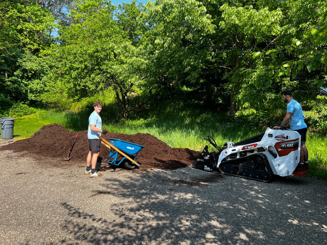 Man using a wheelbarrow next to a landscaping machine and a large pile of mulch outdoors.