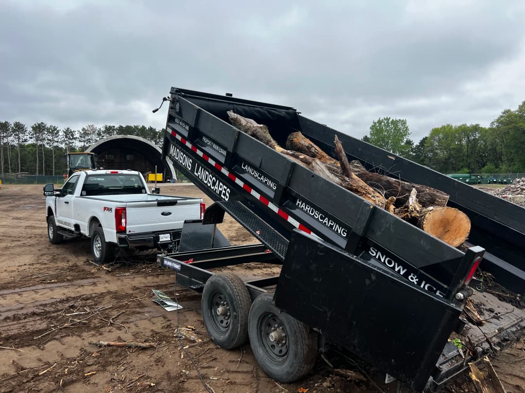 White pickup truck towing a dump trailer loaded with logs at a landscaping site.