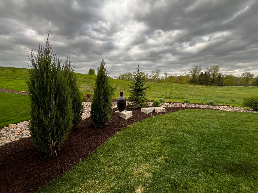 Lush green lawn with decorative stones, black vase, and evergreen shrubs under cloudy sky.