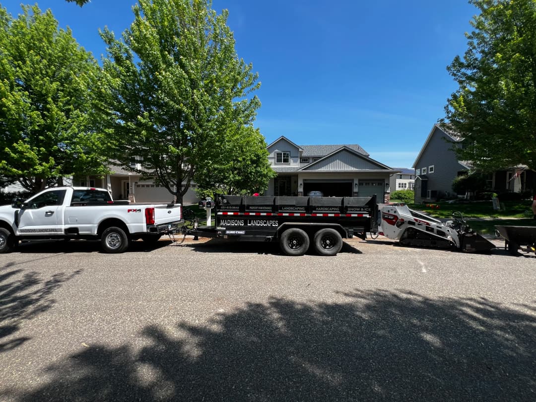 Landscaping truck and trailer parked in residential neighborhood with green trees.