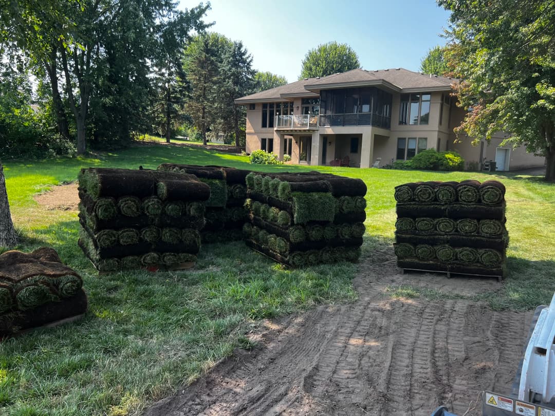 Stacks of rolled sod on a lawn with a house and trees in the background.