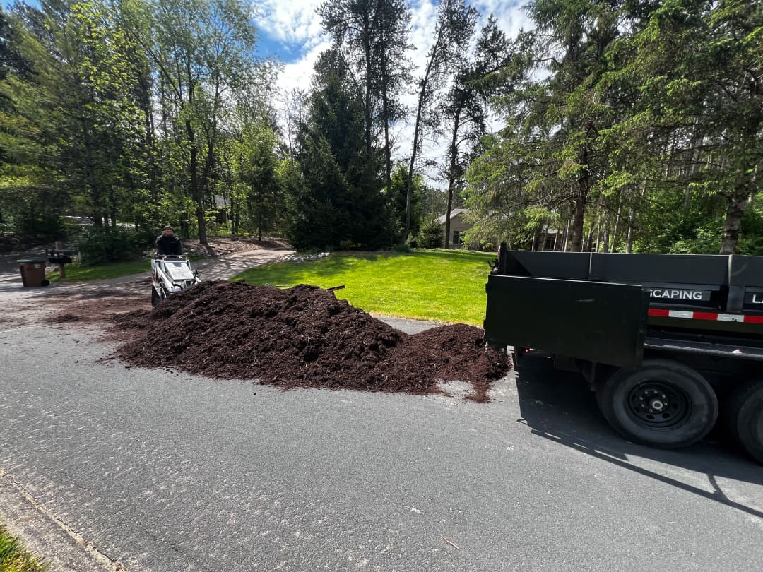 Landsacping scene with a large mulch pile beside a trailer on a residential driveway.