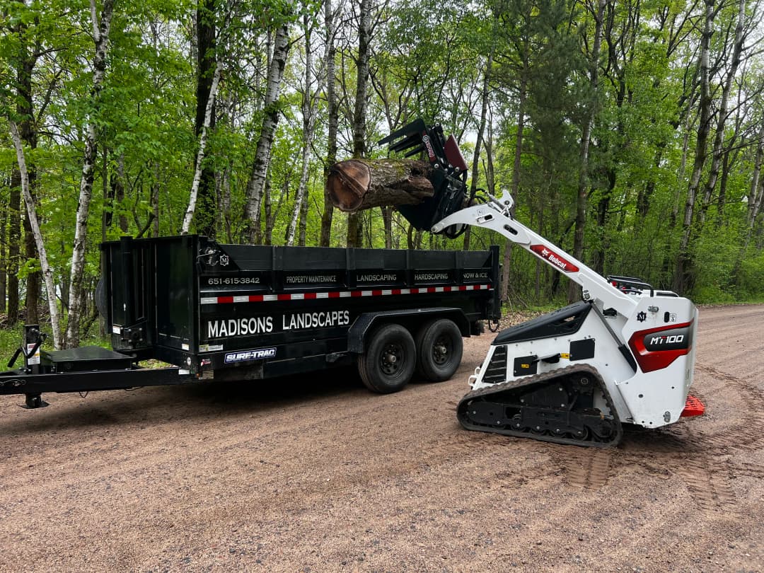 Bobcat HT100 loading a log into a Madison's Landscapes trailer in a wooded area.