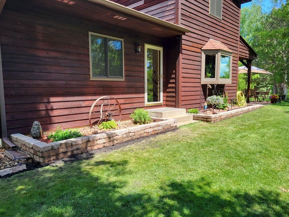 Brown wooden house exterior with landscaped garden, stone borders, and a wheel decoration.