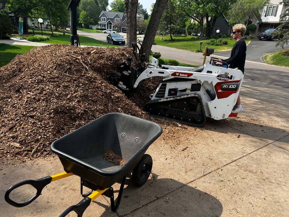 Man operating a Bobcat equipment near a large mulch pile with a wheelbarrow on a driveway.