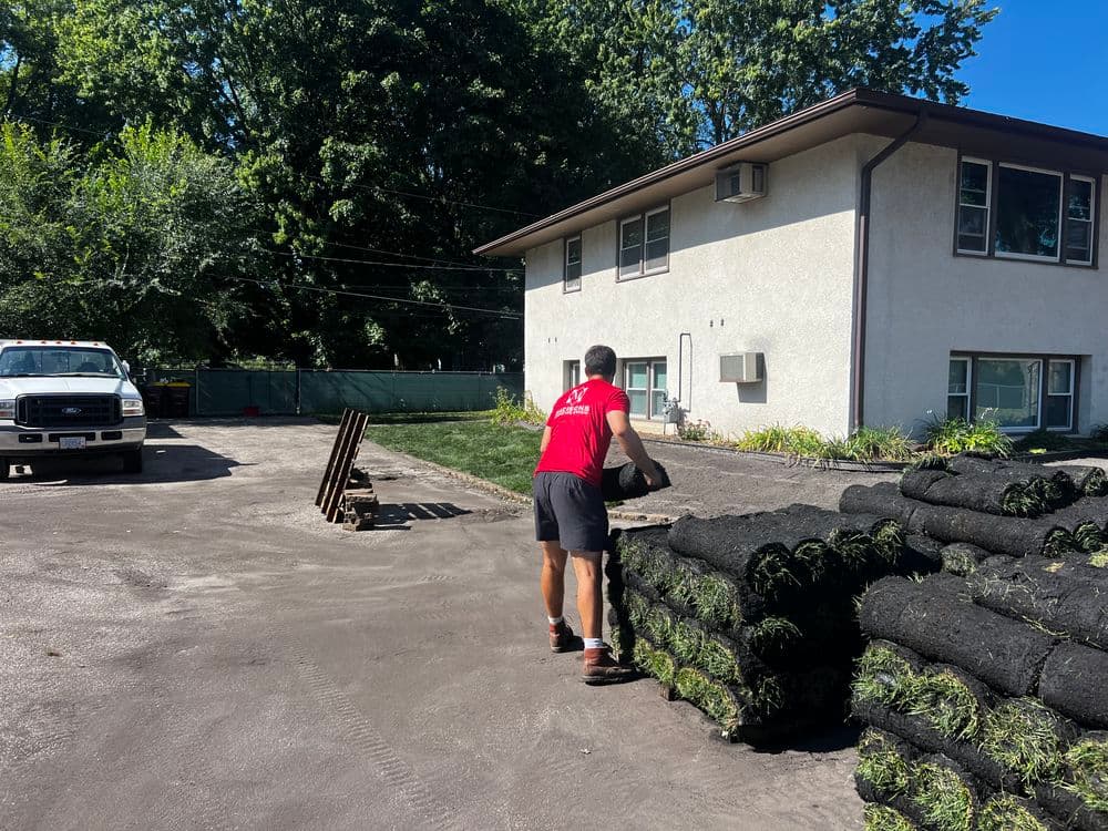 Worker carrying sod rolls to landscaping site beside two-story building.