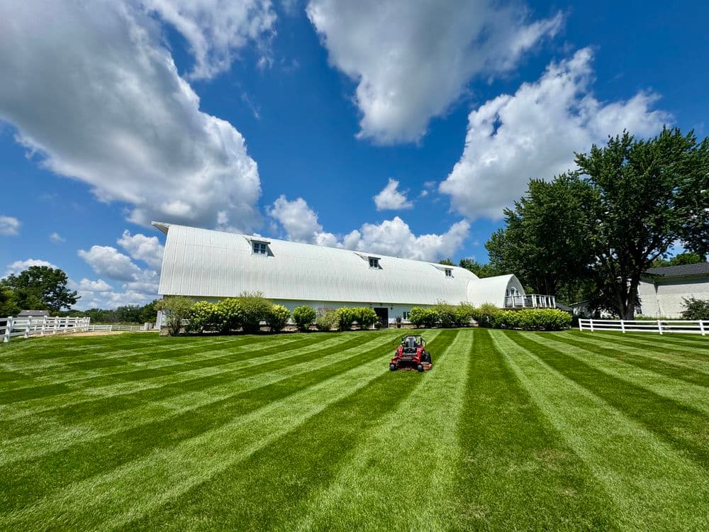 Lawn mower cutting neatly striped grass in front of a modern white barn under a blue sky.