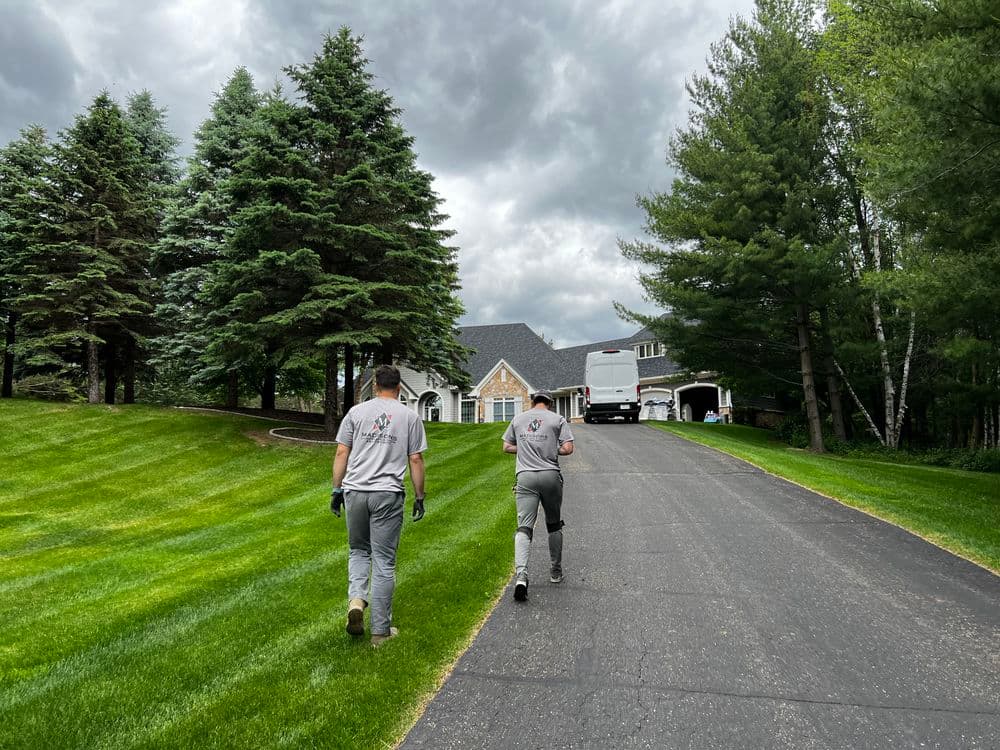 Two men in gray uniforms walking up a paved driveway towards a house surrounded by trees.