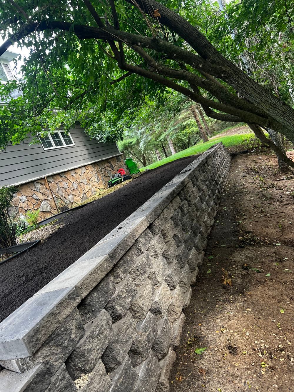 Stone retaining wall with fresh soil, surrounded by trees and a residential backyard.