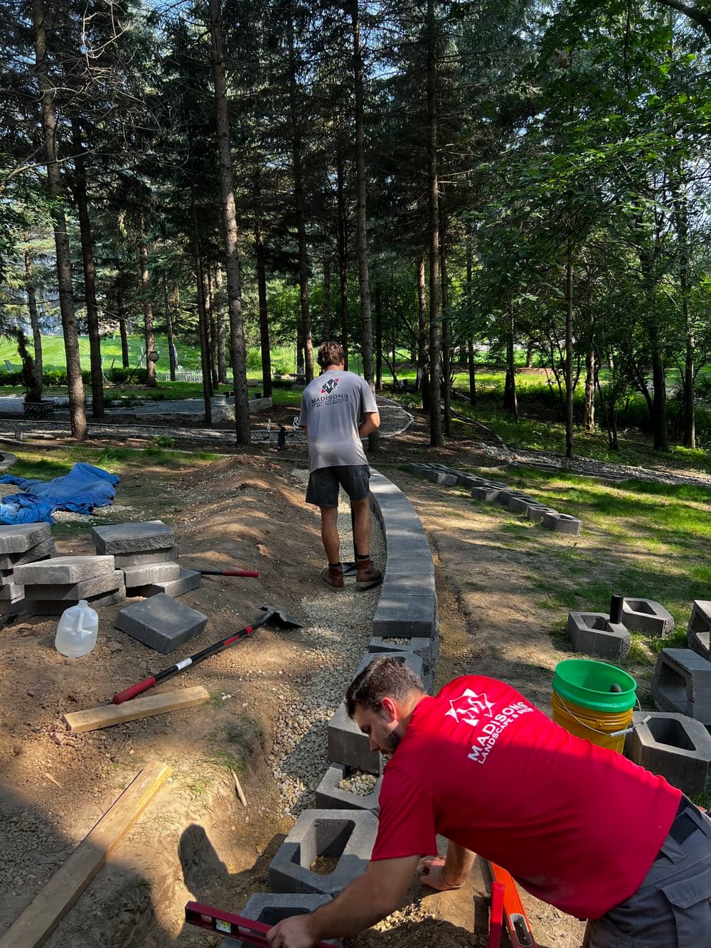 Workers installing landscaping blocks in a wooded area, with tools and materials nearby.