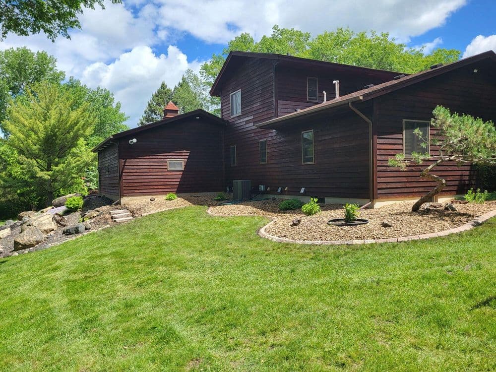 Rustic brown house surrounded by landscaped garden and lush green grass on sunny day.