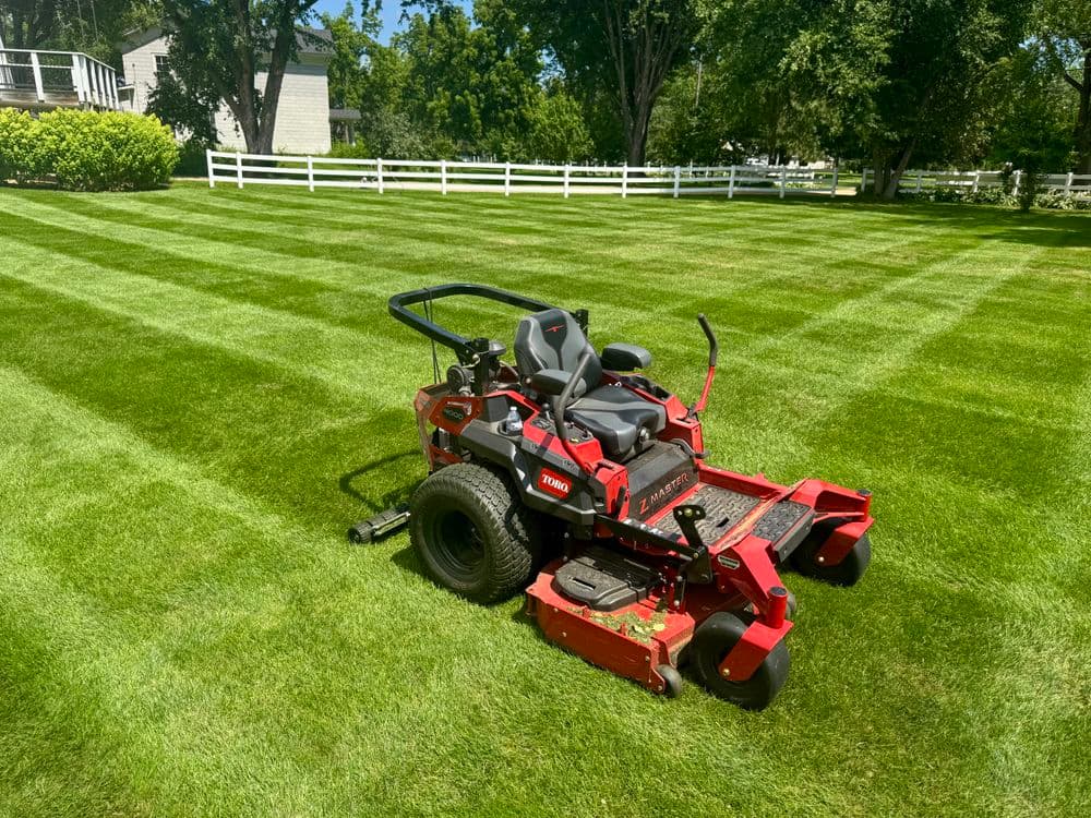 Red riding lawn mower on freshly cut green lawn with neat stripes and white fence in background.