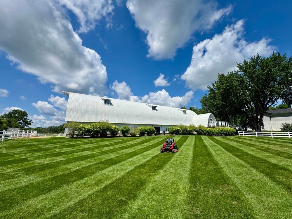 Lawn mower cutting grass near a white barn under a sunny blue sky with fluffy clouds.
