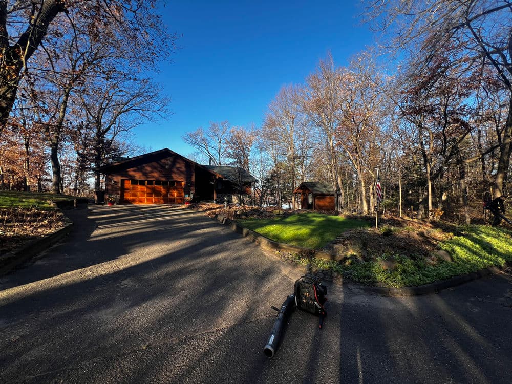 Modern wooden cabin in a wooded area with sunlight casting long shadows on the driveway.